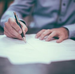 Un homme en chemise à manches longues bleue en train d'écrire sur des feuilles de papier avec un stylo à bille.