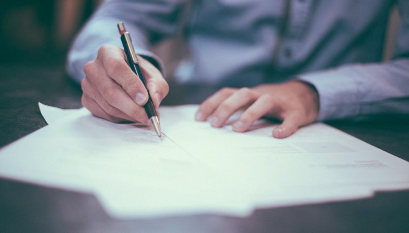 Un homme en chemise à manches longues bleue en train d'écrire sur des feuilles de papier avec un stylo à bille.